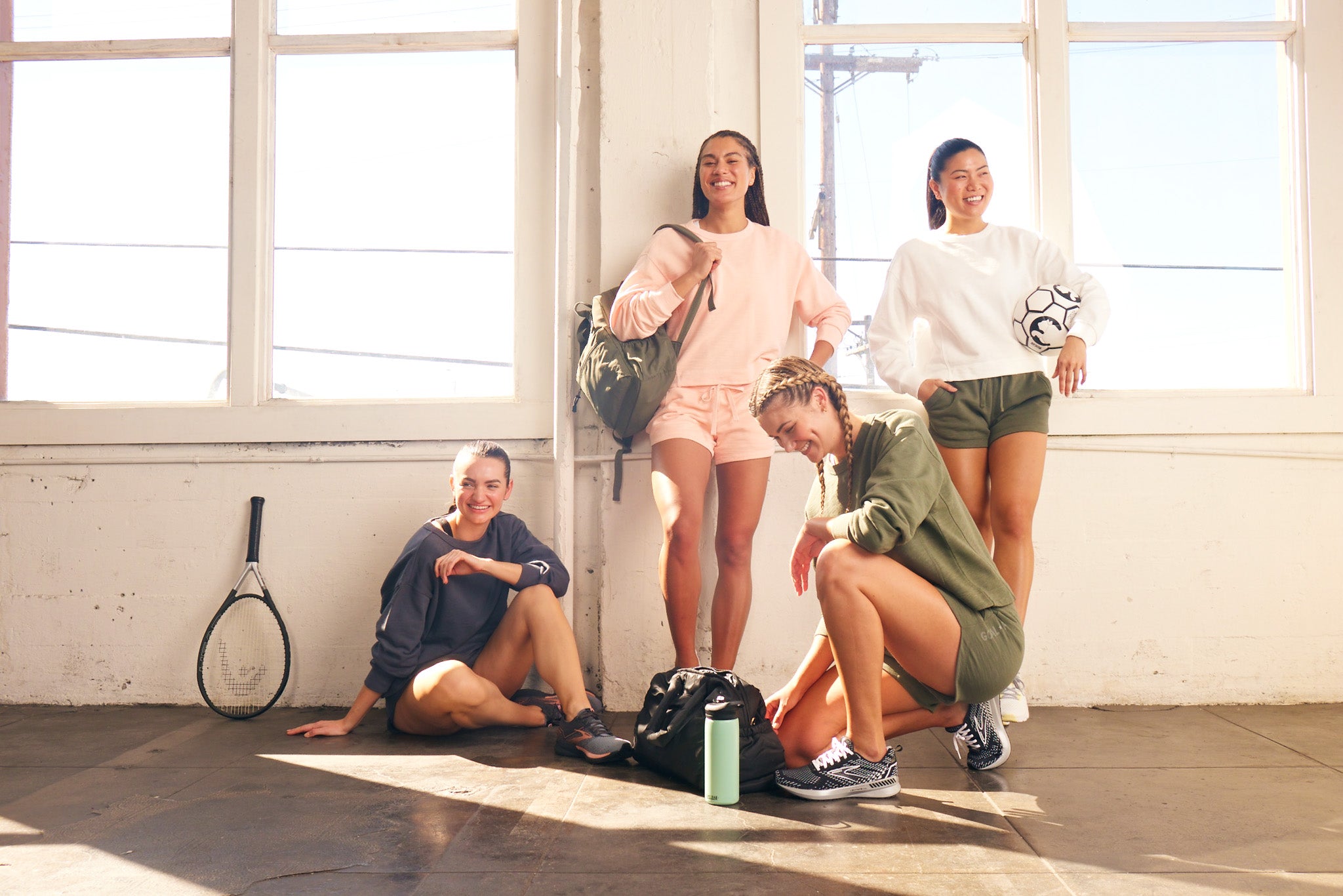 Four young women in athletic wear pose in a sunlit room with sports gear, including a tennis racket, soccer ball, and water bottles, smiling and looking relaxed and happy.