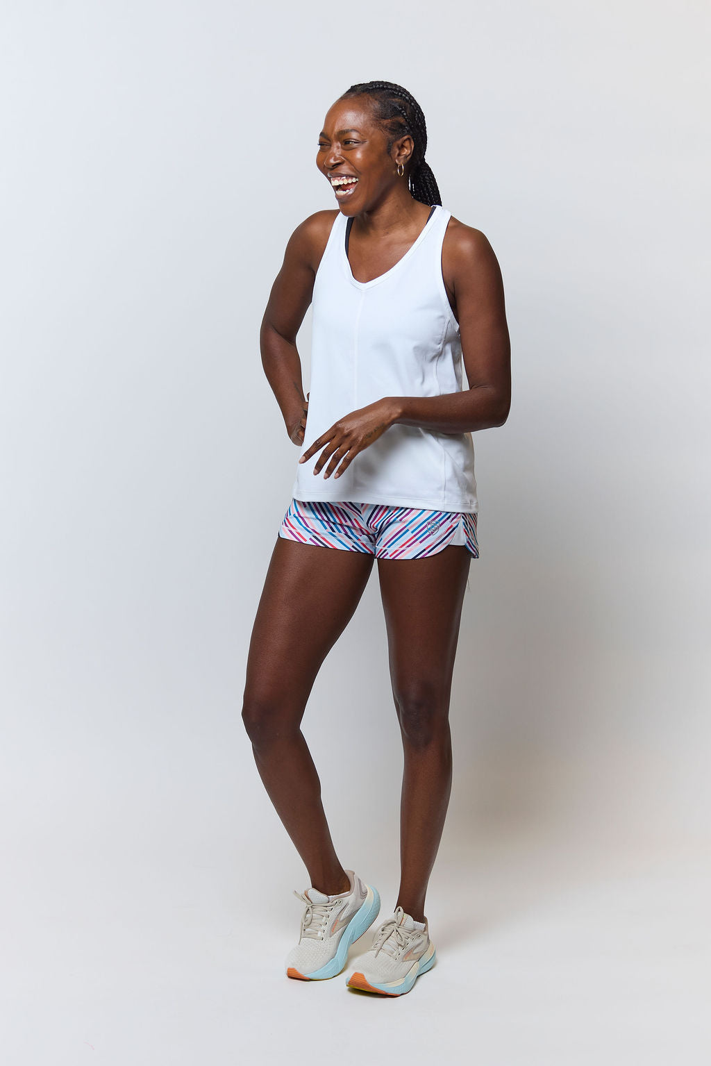 A woman smiles in the Mesh It To Me Tank, featuring sweat-wicking fabric, paired with colorful striped athletic shorts and light running shoes, posed against a plain light background.