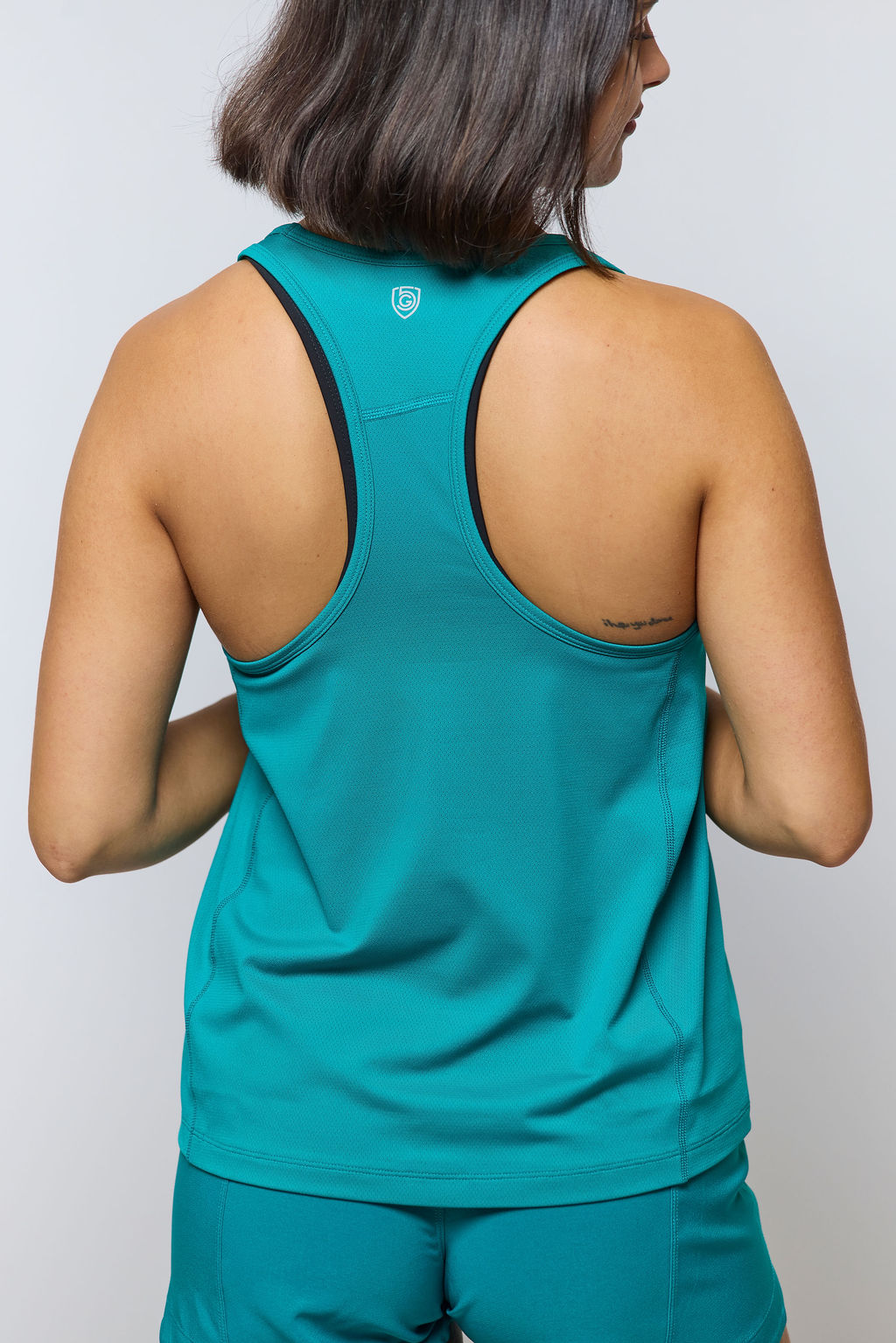 A person with shoulder-length dark hair is shown from the back, wearing the Mesh It To Me Tank and matching shorts. A small tattoo appears on their upper right shoulder.