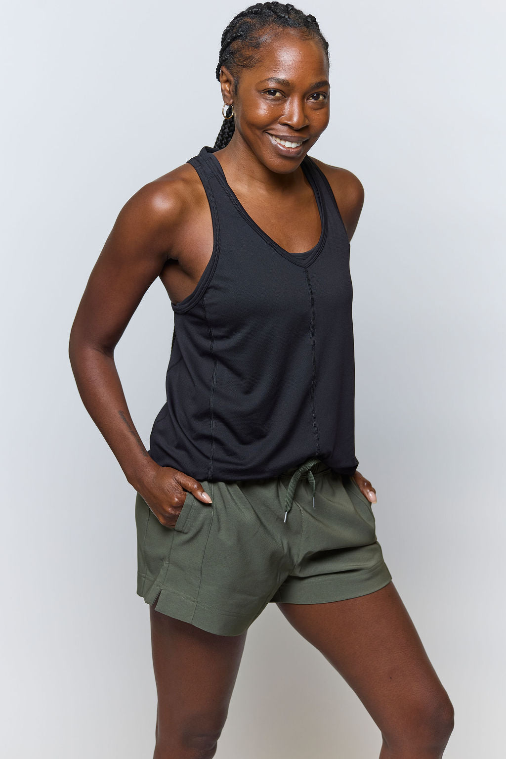 A woman smiles with her hands in the pockets of her olive green shorts, wearing the Mesh It To Me Tank. Her braided hair and a light background emphasize the comfort and sweat-wicking style of this sleek athletic wear.