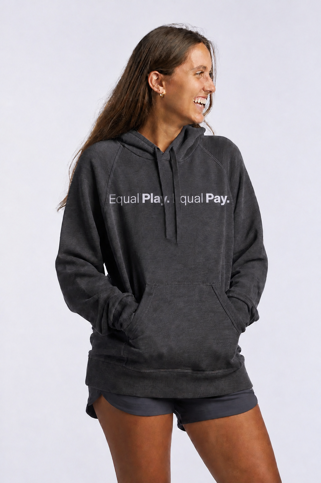 A woman with long brown hair smiles with hands in her pockets, wearing the Equal Play Freasy Hoodie—an ultra-soft dark hoodie featuring “Equal Play. Equal Pay.”—paired with black shorts against a plain light background.