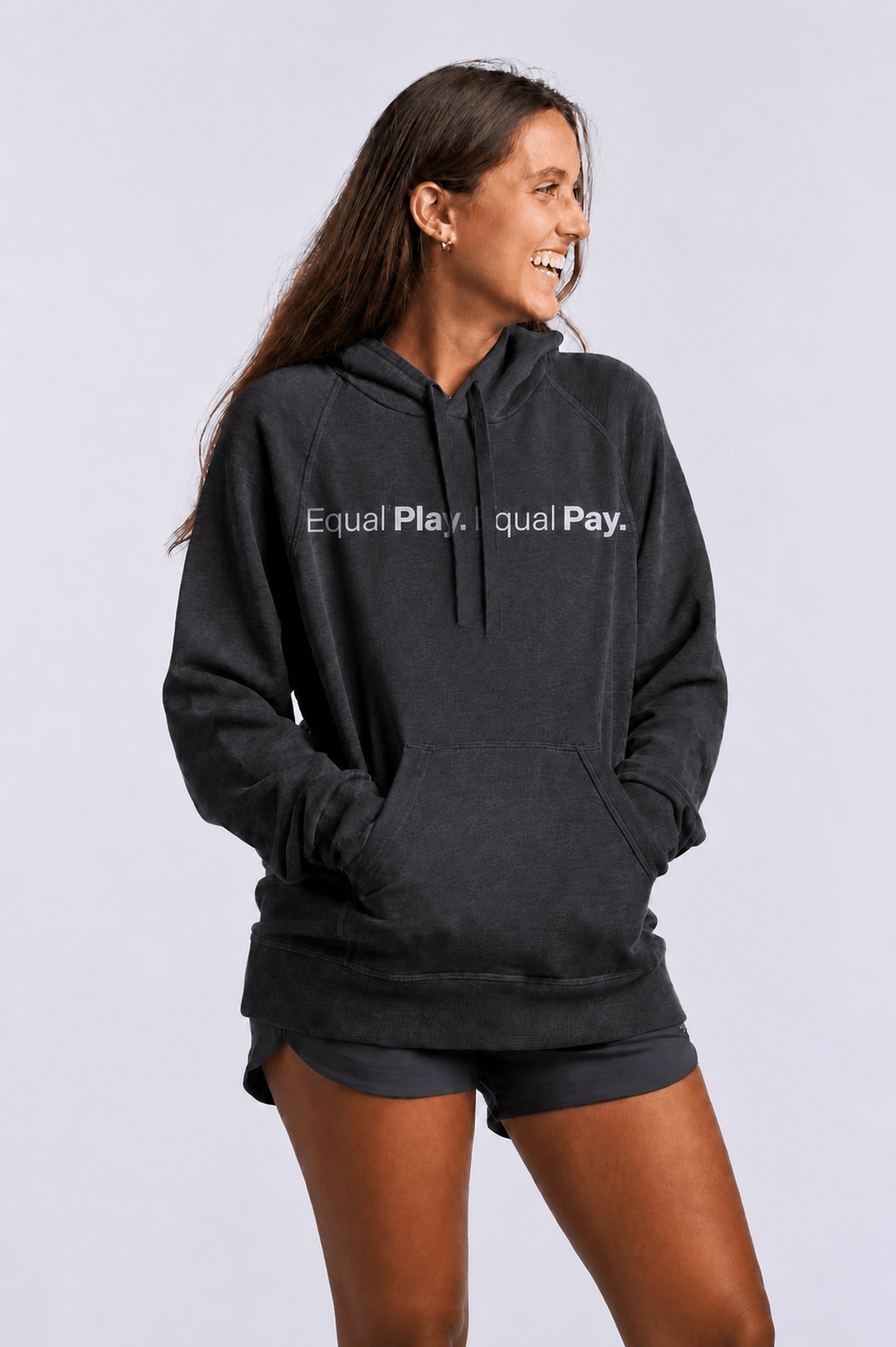 A smiling woman stands with her hands in the pocket of the Equal Play Freasy Hoodie, paired with matching shorts. The ultra-soft black hoodie features “Equal Play. Equal Pay.” on the front, set against a plain, light-colored background.