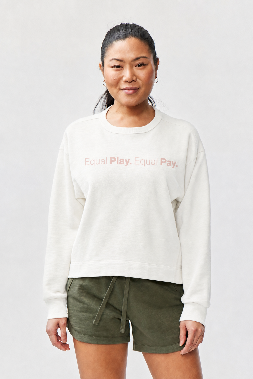 A woman with long dark hair in a ponytail wears the Equal Play Freasy Crew sweatshirt with "Equal Play. Equal Pay." in light pink and olive green shorts, standing against a light gray background, smiling and enjoying its mid weight warmth.