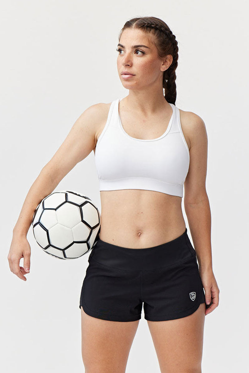 A woman wearing Fast & Free Women's Workout Shorts and a white sports bra holds a soccer ball under her arm, gazing to the side with a focused expression against a plain white background.