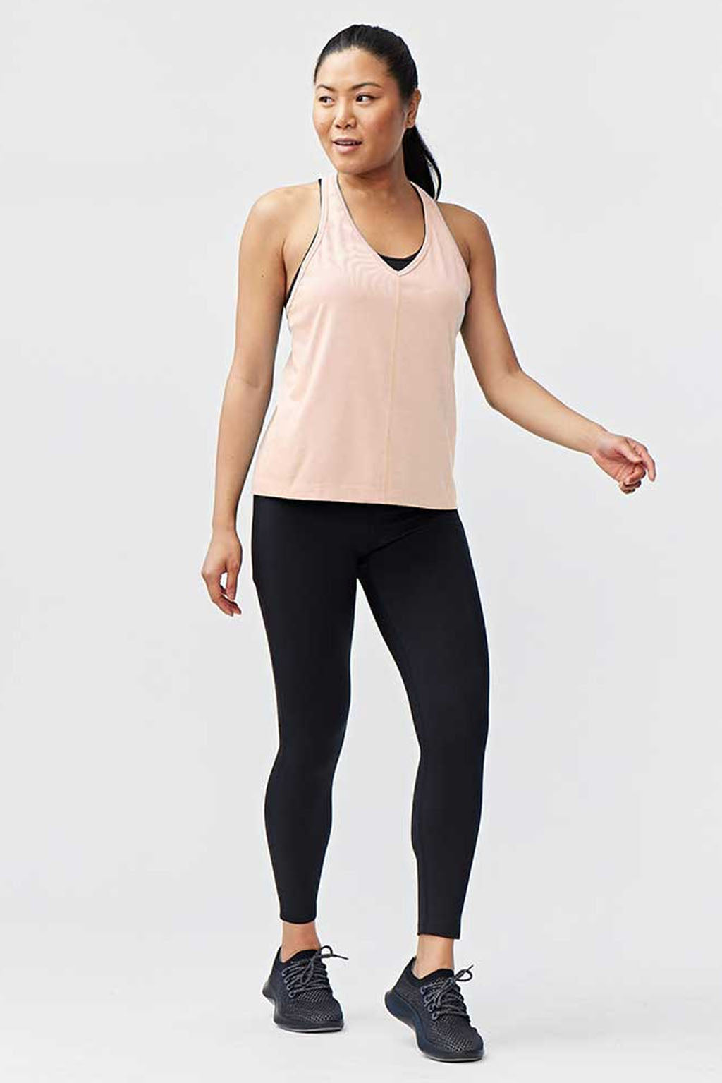A woman with long dark hair tied back wears the Mesh It To Me Tank in light pink, paired with black leggings and shoes, standing against a bright background and looking relaxed to the side.