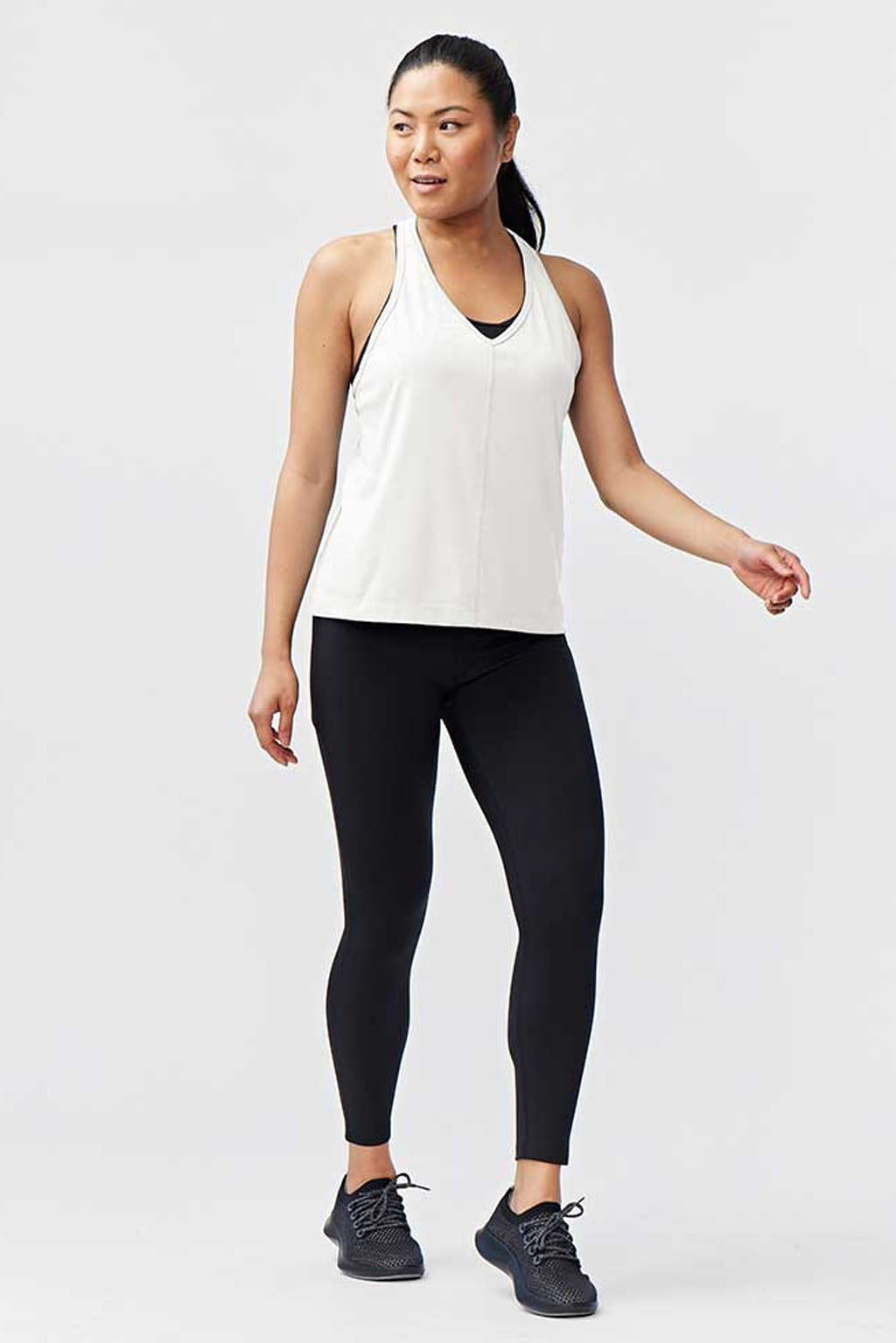 A woman stands against a plain background in the white Mesh It To Me Tank and black leggings with black athletic shoes. Her long dark hair is pulled back as she looks to the side, radiating relaxed confidence in sweat-wicking activewear.