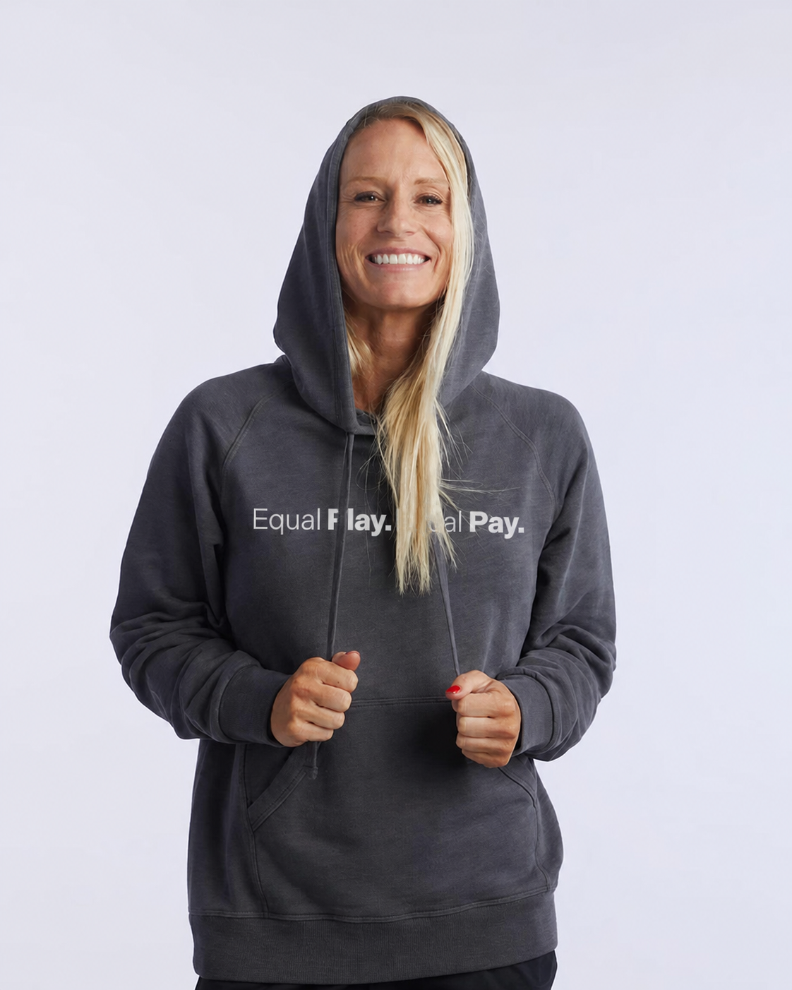 A smiling woman with long blonde hair wears the Equal Play Freasy Hoodie, featuring "Equal Play. Equal Pay." on the front, hood up, standing against a plain light background.