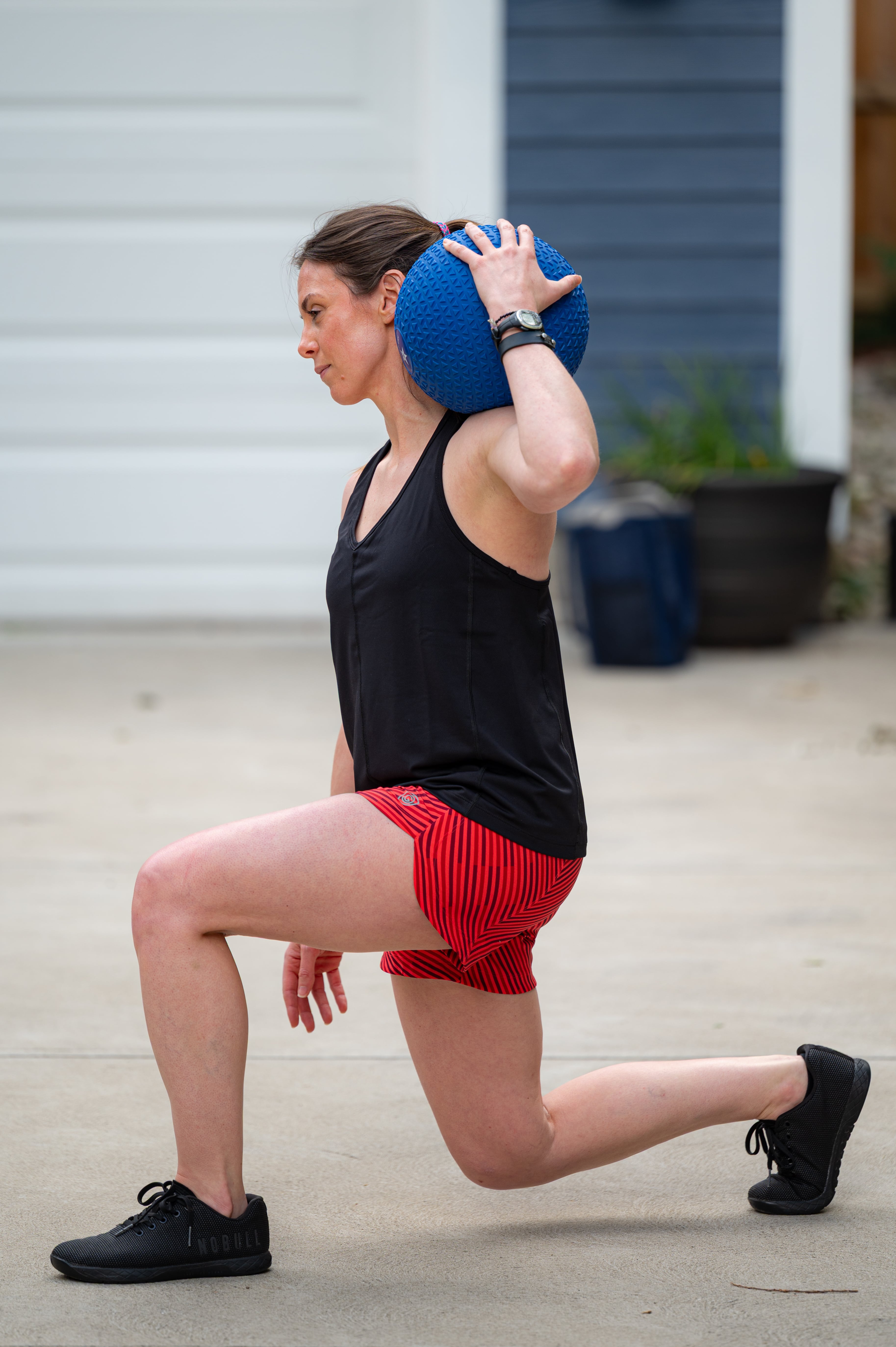 Wearing the Mesh It To Me Tank, a woman does a lunge outdoors with a blue textured ball by her shoulder, paired with red striped shorts and black sneakers for cool, comfortable movement.