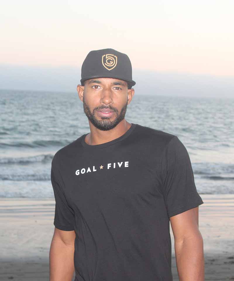 A man walks on the beach in the Goal Five black cotton T shirt for men.