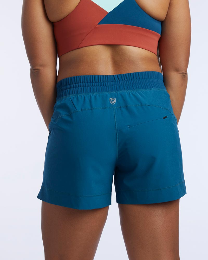 A woman shows the smooth seams and rear zippered pocket of Triple V women's athletic shorts with pockets.