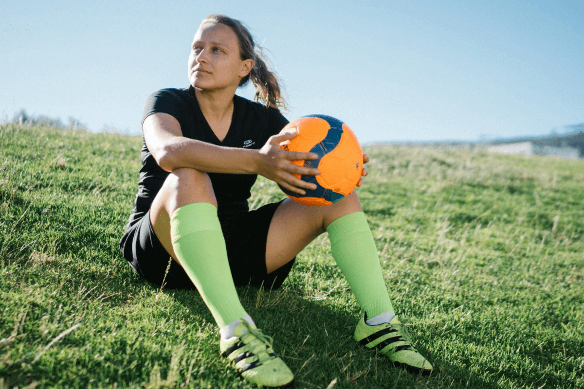 A female soccer player sitting on the side of a hill and holding an orange soccer ball