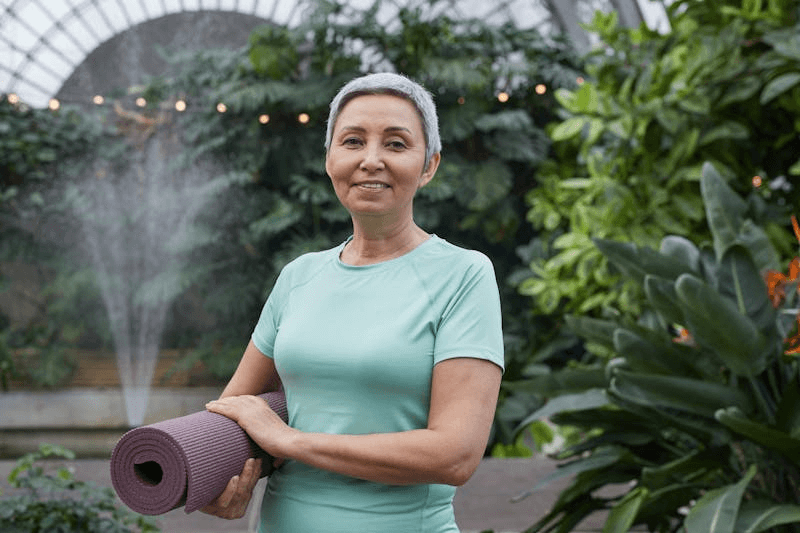 an older woman holding a yoga mat and wearing a workout shirt
