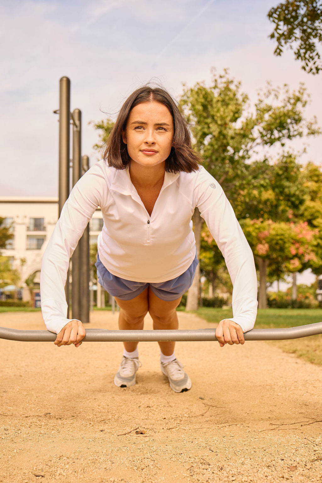 Wearing the Foudy Mid-Weight Long Sleeve ½ Zip Top, a woman does an incline push-up on a metal bar at an outdoor park, with trees and buildings visible behind her.