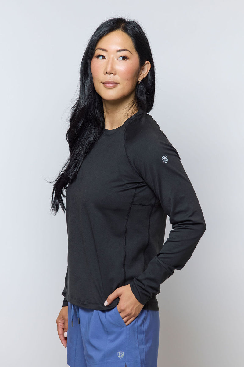 A woman with long black hair stands against a plain background, wearing the Foudy 2.0 Women's Long Sleeve Athletic Top made from moisture-wicking fabric and light blue pants. She poses with one hand in her pocket, looking slightly to the side.