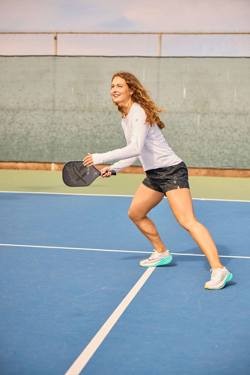 A woman wearing the Indie 2.0 Black Camo shorts and a white Foudy long sleeve top playing pickleball.