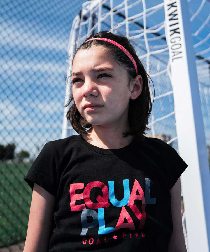 The tween model sits outside featuring the Equal Play black T shirt for girls.